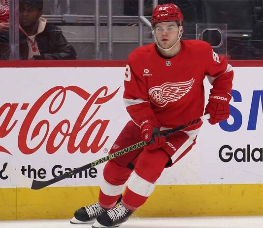 Will the Detroit Red Wings Trade Alex DeBrincat This Offseason? Detroit Red Wings forward Alex DeBrincat skating on the ice, wearing the red winged-wheel jersey, with Coca-Cola and Samsung advertisements on the rink boards.
