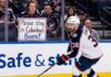 Columbus Blue Jackets captain Boone Jenner skating near the boards during an NHL game while a fan holds a sign reading "Please stay in Columbus Boone!"