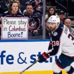 Columbus Blue Jackets captain Boone Jenner skating near the boards during an NHL game while a fan holds a sign reading "Please stay in Columbus Boone!"