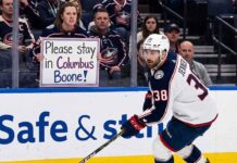 Columbus Blue Jackets captain Boone Jenner skating near the boards during an NHL game while a fan holds a sign reading "Please stay in Columbus Boone!"