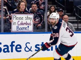 Columbus Blue Jackets captain Boone Jenner skating near the boards during an NHL game while a fan holds a sign reading "Please stay in Columbus Boone!"