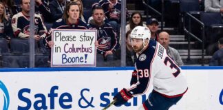 Columbus Blue Jackets captain Boone Jenner skating near the boards during an NHL game while a fan holds a sign reading "Please stay in Columbus Boone!"