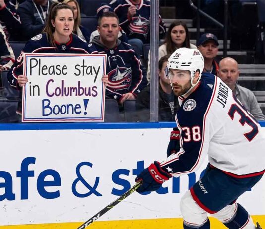 Columbus Blue Jackets captain Boone Jenner skating near the boards during an NHL game while a fan holds a sign reading "Please stay in Columbus Boone!"