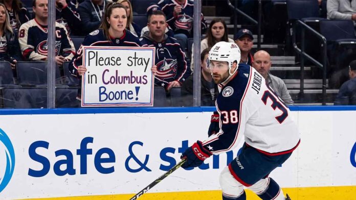 Columbus Blue Jackets captain Boone Jenner skating near the boards during an NHL game while a fan holds a sign reading "Please stay in Columbus Boone!"