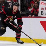 A detailed action photograph of Ottawa Senators captain Brady Tkachuk in his alternate black jersey, skating intensely with a puck on his stick. In the background, behind the glass, a cheerful fan holds up a large, legible sign that says: 'Please stay in Ottawa Brady!'. Canadian Tire and Cortche logos are visible on the boards.