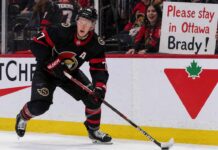 A detailed action photograph of Ottawa Senators captain Brady Tkachuk in his alternate black jersey, skating intensely with a puck on his stick. In the background, behind the glass, a cheerful fan holds up a large, legible sign that says: 'Please stay in Ottawa Brady!'. Canadian Tire and Cortche logos are visible on the boards.