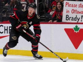 A detailed action photograph of Ottawa Senators captain Brady Tkachuk in his alternate black jersey, skating intensely with a puck on his stick. In the background, behind the glass, a cheerful fan holds up a large, legible sign that says: 'Please stay in Ottawa Brady!'. Canadian Tire and Cortche logos are visible on the boards.