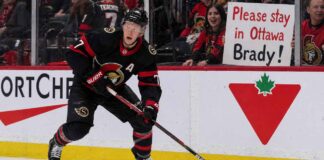 A detailed action photograph of Ottawa Senators captain Brady Tkachuk in his alternate black jersey, skating intensely with a puck on his stick. In the background, behind the glass, a cheerful fan holds up a large, legible sign that says: 'Please stay in Ottawa Brady!'. Canadian Tire and Cortche logos are visible on the boards.
