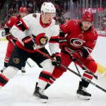 ntense action shot of an Ottawa Senators hockey player (number 7) and a Carolina Hurricanes hockey player (number 71) locked in a close puck battle on the ice during an NHL game, with other players, crowd and arena details in the background.
