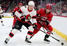 ntense action shot of an Ottawa Senators hockey player (number 7) and a Carolina Hurricanes hockey player (number 71) locked in a close puck battle on the ice during an NHL game, with other players, crowd and arena details in the background.