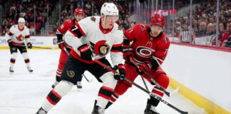 ntense action shot of an Ottawa Senators hockey player (number 7) and a Carolina Hurricanes hockey player (number 71) locked in a close puck battle on the ice during an NHL game, with other players, crowd and arena details in the background.