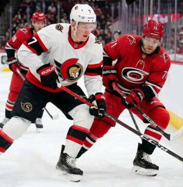 ntense action shot of an Ottawa Senators hockey player (number 7) and a Carolina Hurricanes hockey player (number 71) locked in a close puck battle on the ice during an NHL game, with other players, crowd and arena details in the background.