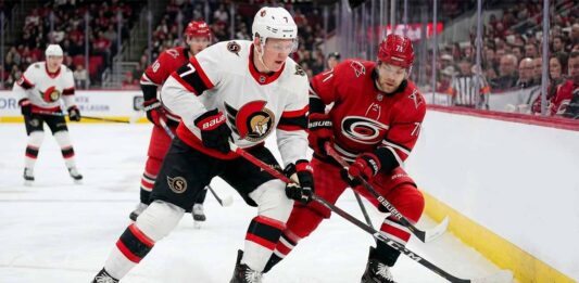 ntense action shot of an Ottawa Senators hockey player (number 7) and a Carolina Hurricanes hockey player (number 71) locked in a close puck battle on the ice during an NHL game, with other players, crowd and arena details in the background.