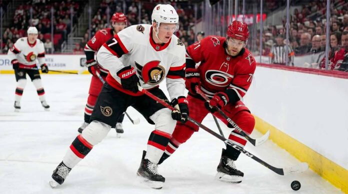 ntense action shot of an Ottawa Senators hockey player (number 7) and a Carolina Hurricanes hockey player (number 71) locked in a close puck battle on the ice during an NHL game, with other players, crowd and arena details in the background.