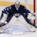 Connor Hellebuyck, star goaltender for the Winnipeg Jets, in focused butterfly stance in full navy blue and white gear in front of the net during an NHL game, amidst recent trade rumors.