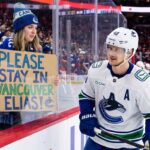 Vancouver Canucks Trade Rumors: Will Elias Pettersson Waive His NMC? A Vancouver Canucks fan holds up a handmade sign behind the glass at Rogers Arena reading "PLEASE STAY IN VANCOUVER ELIAS!" as star center Elias Pettersson (#40) looks on from the ice during an NHL game amidst trade rumors.