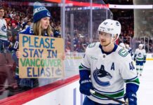 A Vancouver Canucks fan holds up a handmade sign behind the glass at Rogers Arena reading "PLEASE STAY IN VANCOUVER ELIAS!" as star center Elias Pettersson (#40) looks on from the ice during an NHL game amidst trade rumors.
