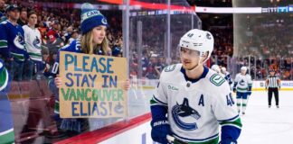 A Vancouver Canucks fan holds up a handmade sign behind the glass at Rogers Arena reading "PLEASE STAY IN VANCOUVER ELIAS!" as star center Elias Pettersson (#40) looks on from the ice during an NHL game amidst trade rumors.