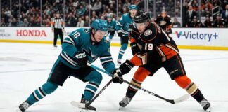 Cutter Gauthier of the Anaheim Ducks, wearing jersey #19, and Macklin Celebrini of the San Jose Sharks, wearing jersey #18, in a face-off battle for the puck on the ice at Honda Center. The arena has Honda Center and Victory+ signage in the background during a San Jose Sharks vs Anaheim Ducks game.