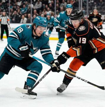Cutter Gauthier of the Anaheim Ducks, wearing jersey #19, and Macklin Celebrini of the San Jose Sharks, wearing jersey #18, in a face-off battle for the puck on the ice at Honda Center. The arena has Honda Center and Victory+ signage in the background during a San Jose Sharks vs Anaheim Ducks game.