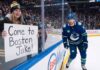 A smiling female fan in a Boston Bruins beanie and jersey sits behind the glass at an NHL game holding a handwritten sign that reads, "Come to Boston Jake!". Beside her, Vancouver Canucks winger Jake DeBrusk skatjes near the boards during play.