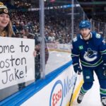 A smiling female fan in a Boston Bruins beanie and jersey sits behind the glass at an NHL game holding a handwritten sign that reads, "Come to Boston Jake!". Beside her, Vancouver Canucks winger Jake DeBrusk skatjes near the boards during play.