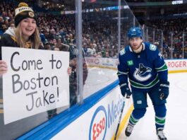 A smiling female fan in a Boston Bruins beanie and jersey sits behind the glass at an NHL game holding a handwritten sign that reads, "Come to Boston Jake!". Beside her, Vancouver Canucks winger Jake DeBrusk skatjes near the boards during play.