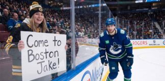 A smiling female fan in a Boston Bruins beanie and jersey sits behind the glass at an NHL game holding a handwritten sign that reads, "Come to Boston Jake!". Beside her, Vancouver Canucks winger Jake DeBrusk skatjes near the boards during play.