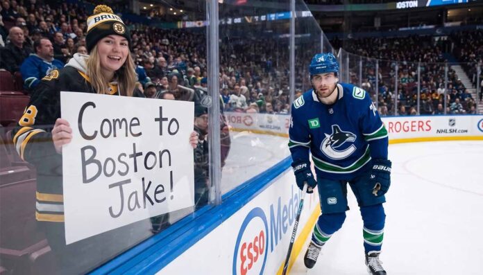 A smiling female fan in a Boston Bruins beanie and jersey sits behind the glass at an NHL game holding a handwritten sign that reads, "Come to Boston Jake!". Beside her, Vancouver Canucks winger Jake DeBrusk skatjes near the boards during play.