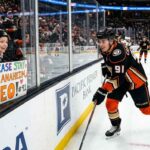 Anaheim Ducks center Leo Carlsson (wearing jersey number 91) smiles at a young female fan in the stands who holds a homemade sign that says, 'PLEASE STAY IN ANAHEIM, LEO! #91'. The image highlights fan anxiety about his potential restricted free agency (RFA) offer sheet status.