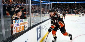 Anaheim Ducks center Leo Carlsson (wearing jersey number 91) smiles at a young female fan in the stands who holds a homemade sign that says, 'PLEASE STAY IN ANAHEIM, LEO! #91'. The image highlights fan anxiety about his potential restricted free agency (RFA) offer sheet status.