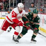 Detroit Red Wings captain Dylan Larkin in a white jersey battles Minnesota Wild forward Kirill Kaprizov in a green jersey for the puck along the boards at Little Caesars Arena.