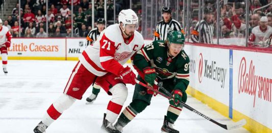 How to Watch Minnesota Wild vs Detroit Red Wings | April 5, 2026 Detroit Red Wings captain Dylan Larkin in a white jersey battles Minnesota Wild forward Kirill Kaprizov in a green jersey for the puck along the boards at Little Caesars Arena.