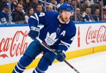 Toronto Maple Leafs defenseman Morgan Rielly #44 skating in an NHL game, holding a hockey stick near the Coca-Cola branded boards.