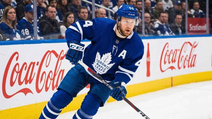 Toronto Maple Leafs defenseman Morgan Rielly #44 skating in an NHL game, holding a hockey stick near the Coca-Cola branded boards.