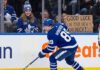An action photograph of Toronto Maple Leafs forward Nick Robertson skating with the puck, while a fan in the stands holds a cardboard sign reading "GOOD LUCK ON YOUR NEW TEAM NICK!" amid NHL trade rumors.
