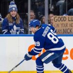 An action photograph of Toronto Maple Leafs forward Nick Robertson skating with the puck, while a fan in the stands holds a cardboard sign reading "GOOD LUCK ON YOUR NEW TEAM NICK!" amid NHL trade rumors.
