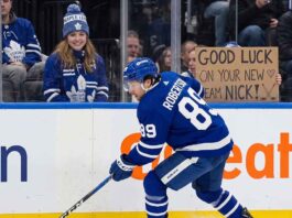 An action photograph of Toronto Maple Leafs forward Nick Robertson skating with the puck, while a fan in the stands holds a cardboard sign reading "GOOD LUCK ON YOUR NEW TEAM NICK!" amid NHL trade rumors.