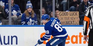 An action photograph of Toronto Maple Leafs forward Nick Robertson skating with the puck, while a fan in the stands holds a cardboard sign reading "GOOD LUCK ON YOUR NEW TEAM NICK!" amid NHL trade rumors.