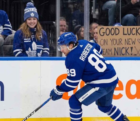 An action photograph of Toronto Maple Leafs forward Nick Robertson skating with the puck, while a fan in the stands holds a cardboard sign reading "GOOD LUCK ON YOUR NEW TEAM NICK!" amid NHL trade rumors.