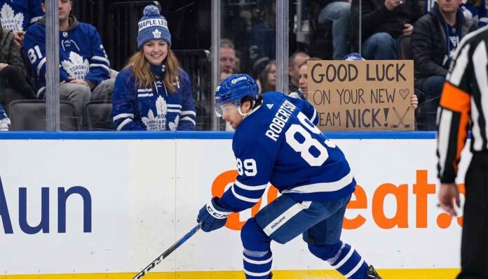 An action photograph of Toronto Maple Leafs forward Nick Robertson skating with the puck, while a fan in the stands holds a cardboard sign reading "GOOD LUCK ON YOUR NEW TEAM NICK!" amid NHL trade rumors.