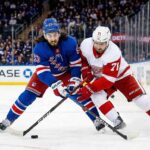 A dynamic photo capturing New York Rangers' Mika Zibanejad and Detroit Red Wings' Dylan Larkin in an intense battle for the puck on the ice at Madison Square Garden during a regular season NHL game. The players are fully engaged, wearing their respective blue home and white away jerseys, with the puck visible between them and a packed arena crowd blurred in the background.
