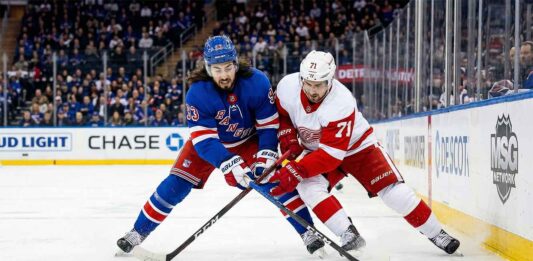 What Channel is the Red Wings vs. Rangers Game On Today? TV & Stream Info A dynamic photo capturing New York Rangers' Mika Zibanejad and Detroit Red Wings' Dylan Larkin in an intense battle for the puck on the ice at Madison Square Garden during a regular season NHL game. The players are fully engaged, wearing their respective blue home and white away jerseys, with the puck visible between them and a packed arena crowd blurred in the background.