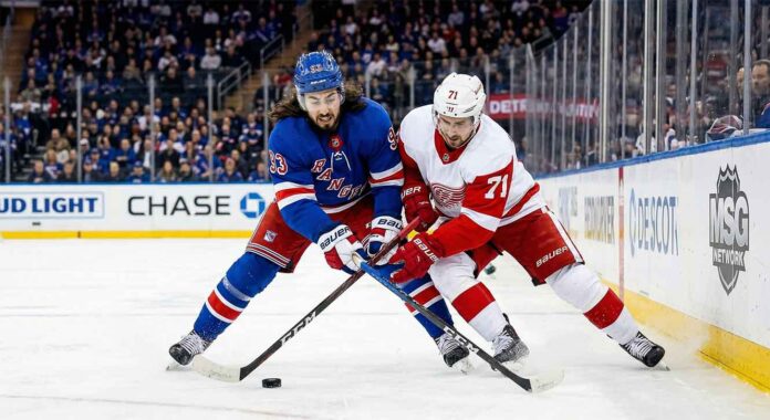 A dynamic photo capturing New York Rangers' Mika Zibanejad and Detroit Red Wings' Dylan Larkin in an intense battle for the puck on the ice at Madison Square Garden during a regular season NHL game. The players are fully engaged, wearing their respective blue home and white away jerseys, with the puck visible between them and a packed arena crowd blurred in the background.