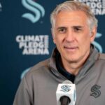 A photograph of Ron Francis, wearing a Seattle Kraken jacket, speaking into a microphone in front of a Climate Pledge Arena press conference backdrop.