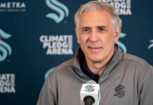 A photograph of Ron Francis, wearing a Seattle Kraken jacket, speaking into a microphone in front of a Climate Pledge Arena press conference backdrop.
