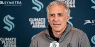 A photograph of Ron Francis, wearing a Seattle Kraken jacket, speaking into a microphone in front of a Climate Pledge Arena press conference backdrop.