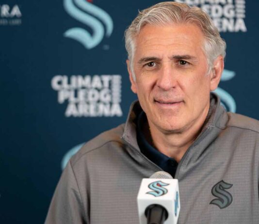 A photograph of Ron Francis, wearing a Seattle Kraken jacket, speaking into a microphone in front of a Climate Pledge Arena press conference backdrop.