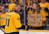 Steven Stamkos of the Nashville Predators in a yellow jersey looks at a female fan holding a cardboard sign that says 'Please don't go Steven!' amidst offseason trade rumors.