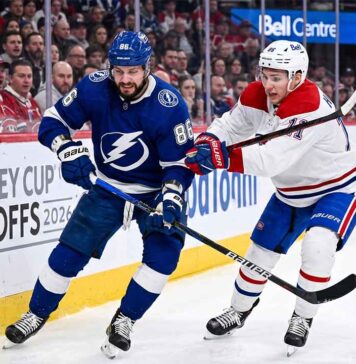 Dynamic on-ice action photo from Game 4 of the 2026 Stanley Cup Playoffs, showing Tampa Bay Lightning forward Nikita Kucherov (#86, blue jersey) battling Montreal Canadiens defenseman Lane Hutson (#?, white jersey) for puck possession near the boards in a packed Bell Centre.